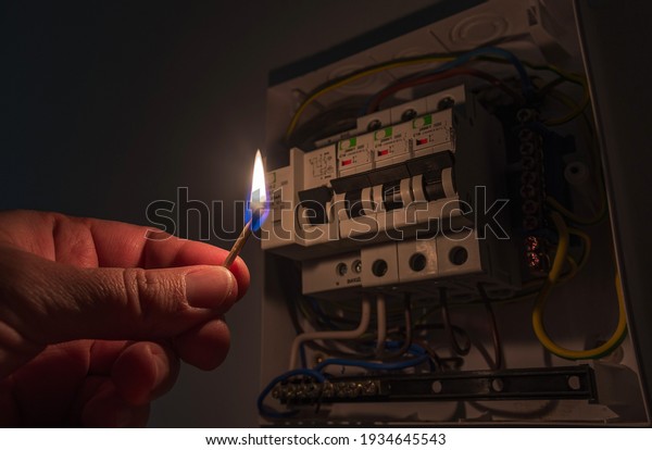 Blackout concept. Person\'s hand in complete\
darkness holding a burning match to investigate a home fuse box\
during a power outage.