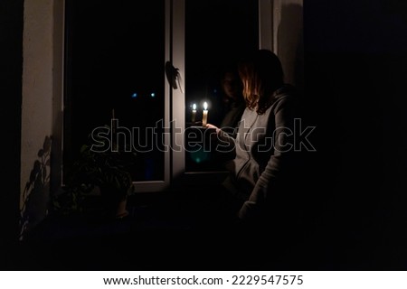 Blackout. Energy crisis. Destruction of infrastructure. Power outage concept. Girl with a burning candle in a dark room sits near the window