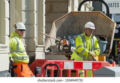 Blackpool, Lancashire/UK - July 18th 2019: male caucasian construction workers wearing high visibility jackets taking a break with road barriers and a digger and stone building in the background