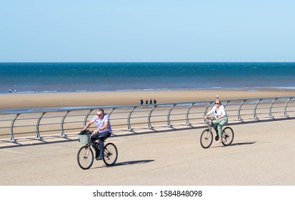 Blackpool, Lancashire/UK - September 21st 2019: Caucasian Man And Woman Cycling Along The Seafront Promenade At Blackpool With People On The Sandy Beach With Sea And Blue Sky Background