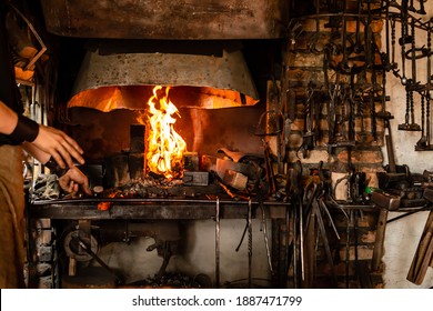 The Blacksmith Holds Billet Over Hot Coals In Clay Oven. Blacksmith Heating Iron Metal Sword Manufacturing Marching Forge, Hands Of A Blacksmith Hold Piece Of Steel In Fire Of A Red-hot Forge