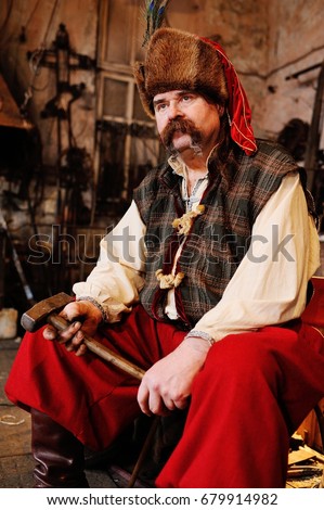 Blacksmith Kozak or a Cossack in Ukrainian national clothes with a hammer in the hands on the background of a blacksmith shop
