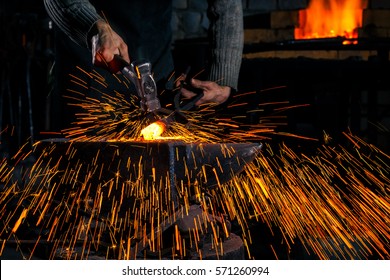 The blacksmith manually forging the molten metal on the anvil in smithy with spark fireworks
