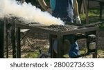 A blacksmith stands beside a coal forge as thick white smoke rises from the fire bed and tools rest on the tray.