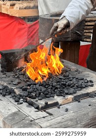 Blacksmith working at a traditional forge. Gloved hand holding metal rod over fire with burning coal. Intense flames, black anvil, rustic wooden workbench. Historical craftsmanship