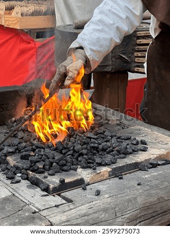 Blacksmith working at a traditional forge. Gloved hand holding metal rod over fire with burning coal. Intense flames, black anvil, rustic wooden workbench. Historical craftsmanship.