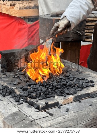 Blacksmith working at a traditional forge. Gloved hand holding metal rod over fire with burning coal. Intense flames, black anvil, rustic wooden workbench. Historical craftsmanship