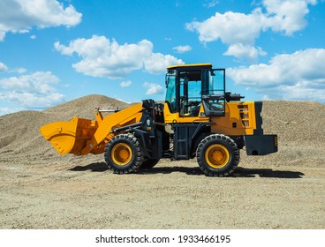 Black-yellow front loader with small wheels against the background of a large pile of stone sand and a blue sky with white clouds. Side view.
