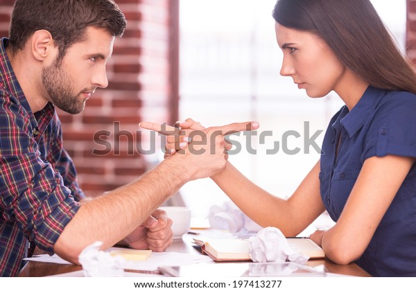 Blaming\
each other. Side view image of angry man and woman sitting face to\
face at the office table and pointing each\
other