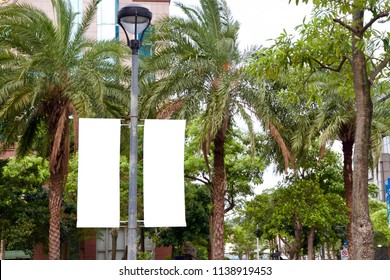 The Blank Advertising Banner Suspended On The Street Lamp Pole With The Tree And Building Facade Background.