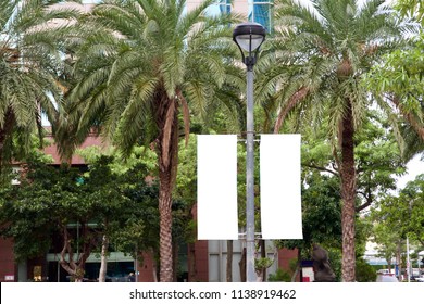 The Blank Advertising Banner Suspended On The Street Lamp Pole With The Tree And Building Facade Background.