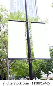 The Blank Advertising Banner Suspended On The Street Lamp Pole With The Tree And Building Facade Background.