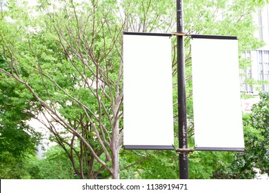 The Blank Advertising Banner Suspended On The Street Lamp Pole With The Tree And Building Facade Background.