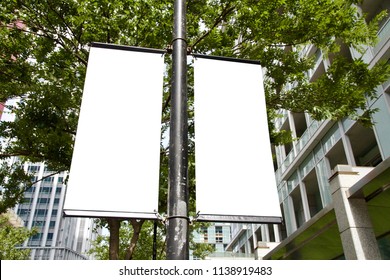 The Blank Advertising Banner Suspended On The Street Lamp Pole With The Tree And Building Facade Background.