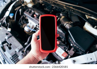 Blank black screen of OBD2 scanner tool in a mechanic hand with a car engine compartment blurred on background, Car engine scanning with wireless technical tool, Car maintenance service concept