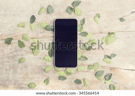 Blank black smartphone screen with leaves on wooden table, mock up