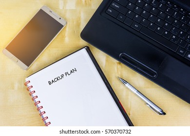 A Blank Book With Text Backup Plan, Smartphone And Laptop.