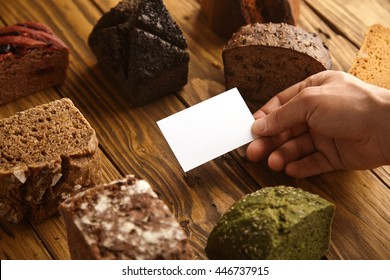Blank business card of professional artisan baker presented in hand in center of many mixed alternative baked exotic bread samples above wooden rustic table - Powered by Shutterstock