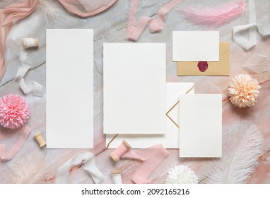 Blank cards and envelopes between pastel flowers, silk ribbons and feathers on marble top view. Romantic scene with wedding suite mockups flat lay,  place for text. Valentines, Spring or romantic 
