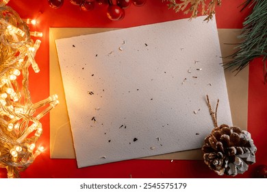 A blank Christmas card rests on a red background surrounded by festive elements like a pinecone, fir branches, and warm string lights, evoking a cozy holiday atmosphere. - Powered by Shutterstock