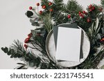 Blank Christmas greeting card, invitation and envelope on plate.  Floral garland of fir, pine tree branches. Olive, eucalyptus leaves with red berries. White table background. Moody flat lay, top view