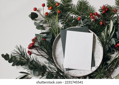 Blank Christmas greeting card, invitation and envelope on plate.  Floral garland of fir, pine tree branches. Olive, eucalyptus leaves with red berries. White table background. Moody flat lay, top view - Powered by Shutterstock