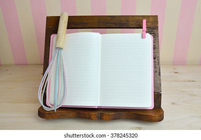 Blank Cook Book And Egg Whisk On Wooden Book Holder With Pink-yellow Background