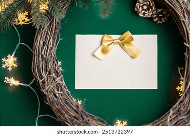 Blank empty Christmas card with a yellow gold ribbon bow on a natural wooden grapevine wreath frame for Christmas decorations with soft snowflake string lights, pine cones, green fabric background DIY - Powered by Shutterstock