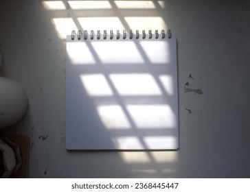 The blank empty page of the opened spiral-spine vintage square notebook is isolated on a textured white desk, under the styled sunlight , for art drawing, text, word, sketch, logo design mockup.