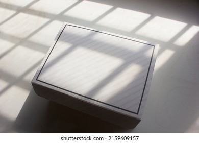 A Blank Empty White Bakery Box With Minimal Stripe Line Is Isolated On A Texture White Table Background, Under The Sunlight Window Shading, For Template Dessert Packaging Design Mockup.