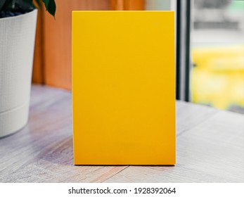 Blank Empty Yellow Booklet, Menu, Paper Stands On Wooden Table In Corner Of Cafe By The Window. Template