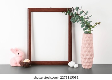 Blank frame, vase with eucalyptus, Easter eggs and rabbit on table near white wall