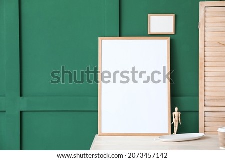 Blank frames and wooden mannequin on table near green wall