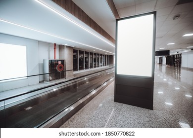 A Blank Glowing Vertical Ver Ad Billboard Placeholder Mock-up Near A Long Travelator Indoors Of A Modern Airport Corridor; A Template Of An Empty White Informational Poster Stand Near A Moving Walkway