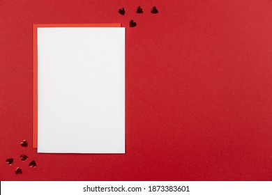 Blank greeting card mockup and envelope on red background with hearts confetti. Valentines, mothers day or wedding flat lay composition. Top view. Copy space. Love concept. 