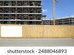 Blank information board on fence outside the construction site with a new large building among the scaffolding, Tower crane and sky. Background for copy space.