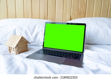 Blank Laptop With Empty Screen And Little Wooden House Lying On The White Bed.