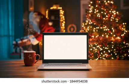 Blank Laptop On A Desktop At Home And Santa Claus Bringing Gifts In The Background, Holidays And Christmas Concept