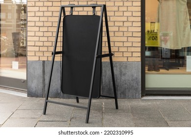 Blank Metal Sandwich Placard Board In Front Of A Window Store On The Street. Black New Modern Signboard For Commercial Business. Or Retail  In The City Center. Copy Space, Empty Space For Advertising