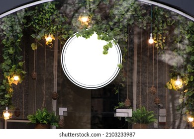 Blank Minimal Circular Shop Signboard Mockup For Design. Street Hanging Sign Board For Logo Presentation With Light Garland Amond The Leaves. Metal Cafe Restaurant Or Bar Badge Black White Round.