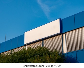 Blank name board on a building exterior. Empty white space for a company logo on a wall. Template of a signboard on an office house. Blue facade with closed window blinds. Blue sky in the background. - Shutterstock ID 2184674799