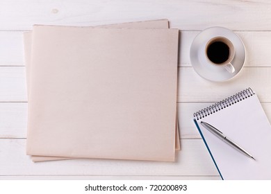 Blank Newspaper On White Wooden Table. Top View Of Daily News Mock Up, Cup Of Coffee, Note Pad And Pen. Space For Your Text Or Image.