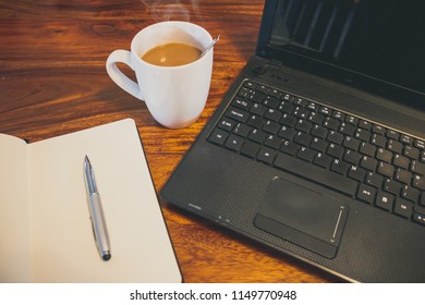 A Blank Note Pad, Silver Metal Pen, Hot Cup Of Coffee In A White Cup With Steam And A Black Computer. All Objects Sitting On A Solid Oak Surface. Shot Taken From Front Eye Level.