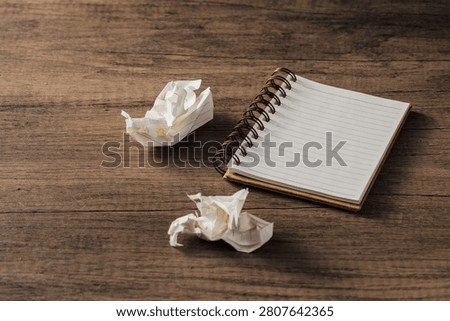 A blank notebook sits on a rustic wooden table next to crumpled paper pieces, symbolizing creativity and the journey from idea to execution in a workspace.