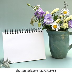 Blank notebook with whitespace beside vase of pretty flowers on green background
				