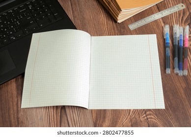 a blank open school squared notebook lies on a brown wooden table near the edge of a black laptop keyboard a metal ruler and four multicolored pens lined up in a row