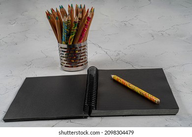 Blank and opened black notepad and a colorful pencil on a wooden surface and lots of colorfol pencils on a metalic pencilcase
