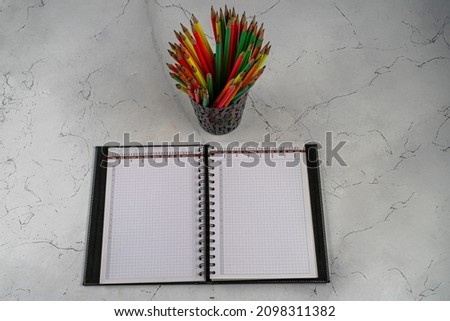 Blank and opened notepad and a colorful pencil on a wooden surface and lots of colorfol pencils on a metalic pencilcase