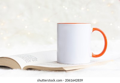 Blank Orange Handle Mug On The Book  With White Bokeh Lights. 11 Oz White Ceramic Mug Mock Up Photo.