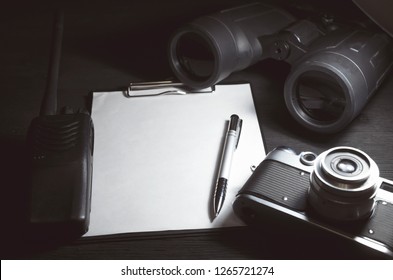 Blank Page With Copy Space, Pen, Portable Radio, Binoculars And Photo Camera On A Black Spy Agent Table Background. Top Secret Documents. Detective Agent Dossier. Interrogation Report.